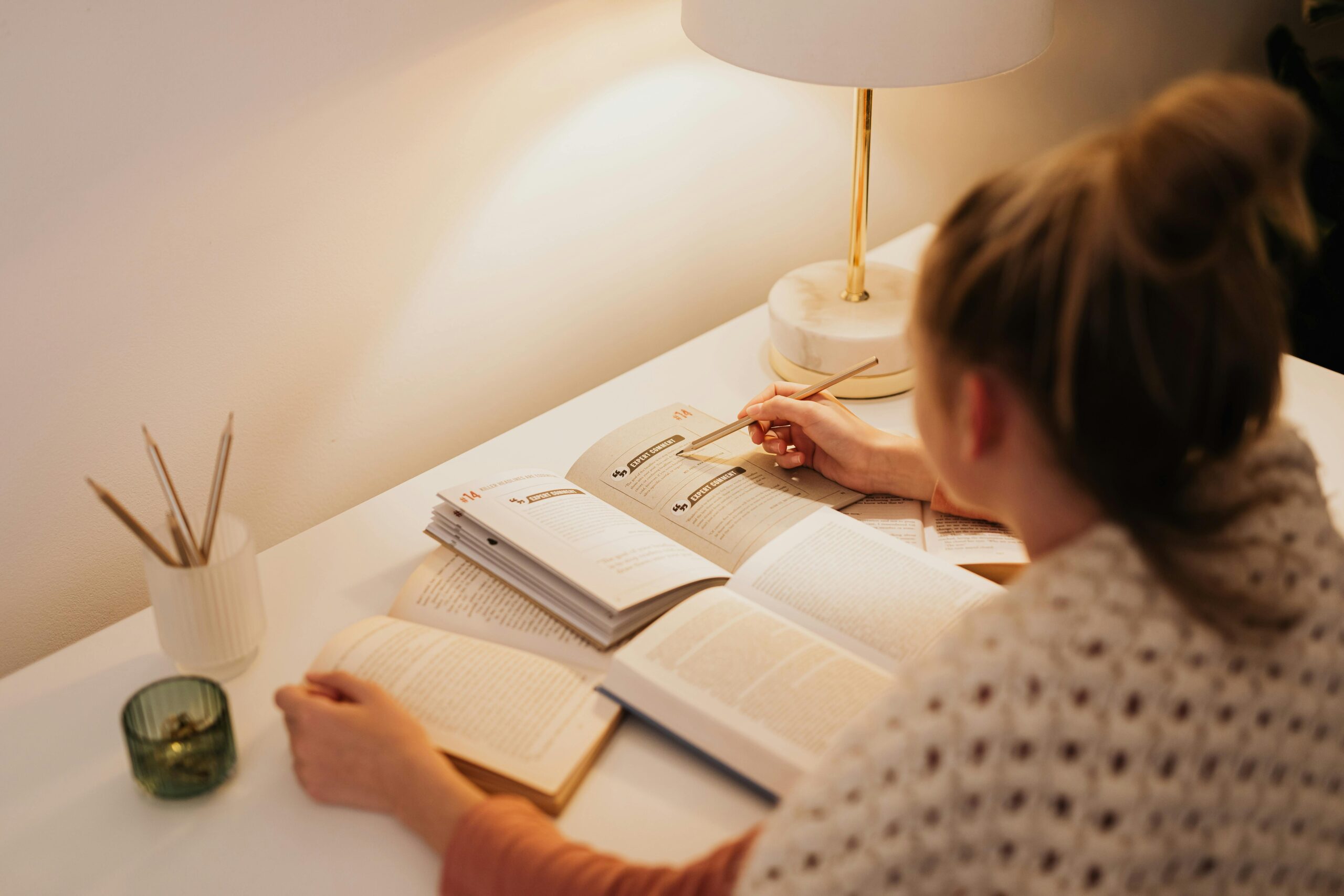 A woman reading and taking notes with books on a desk under a lamp.